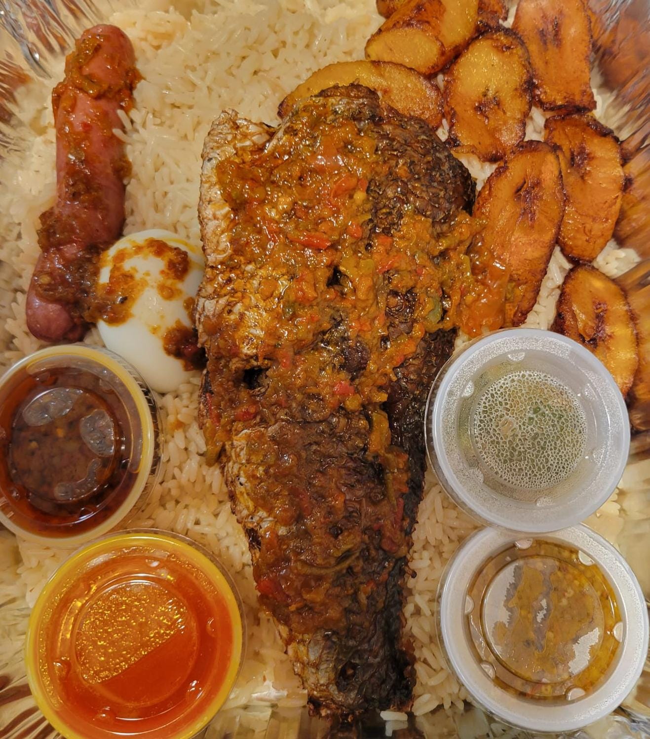 Dry rice and fried fish with plantains, boiled egg, sausage, and sauces